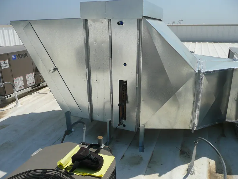 A new galvanized air handler on a commercial roof, its copper coil connections showing, with a yellow legal pad and a camera bag left on the condenser in front.